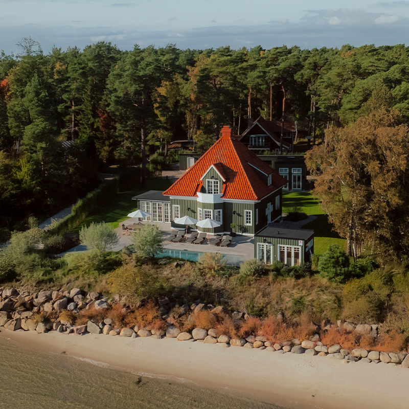 Perfect Hideaways - An elegant forest-side villa with a striking red tiled roof, sage-green wooden cladding, and expansive terraces featuring sun loungers and white umbrellas overlooking a tranquil pool, set among lush greenery. Villa Sand, Ahus