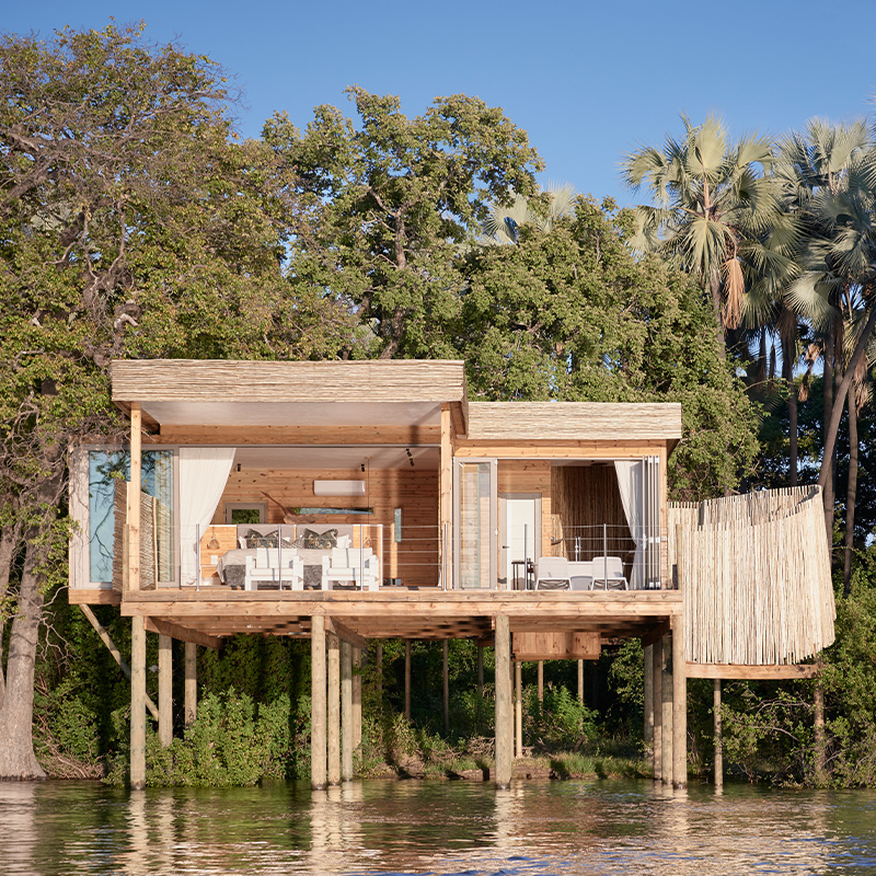 Perfect Hideaways: A luxurious wooden safari lodge elevated on stilts above the Zambezi River, blending seamlessly with the lush riverine forest and palm trees, offering an open-plan design with elegant furnishings and a private deck. Victoria Falls Island Lodge, Zambezi National Park.