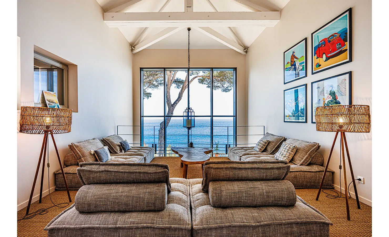 Perfect Hideaways: A cosy lounge with deep cushioned sofas facing a large picture window framing the blue sea and a tall pine tree, accented by woven floor lamps and colourful retro artwork on the wall — Cassis de Luxe, Cassis