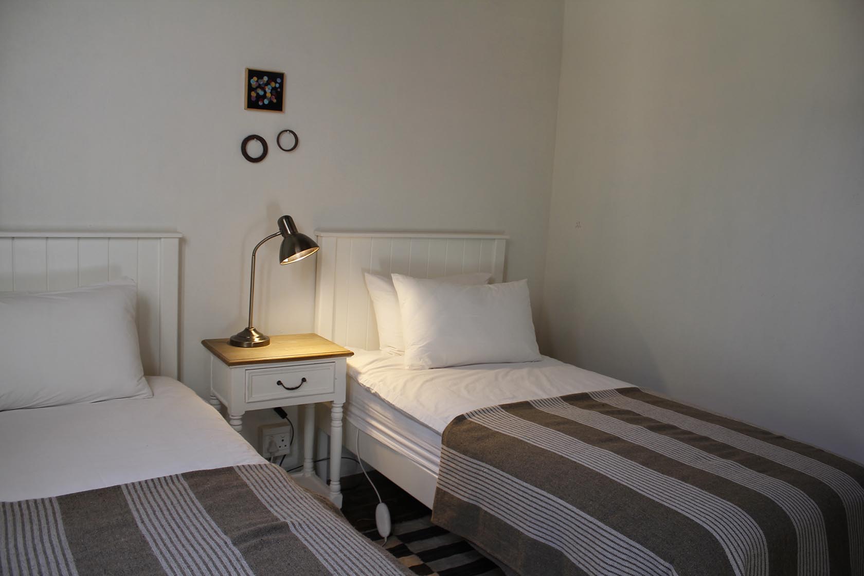 Perfect Hideaways- A simple twin bedroom with two single beds dressed in white linens and striped blankets, centered around a small wooden bedside table with a brass reading lamp and minimalist wall décor above. Karoo Cottage, Sneeuberg Nature Reserve, Eastern Cape, South Africa