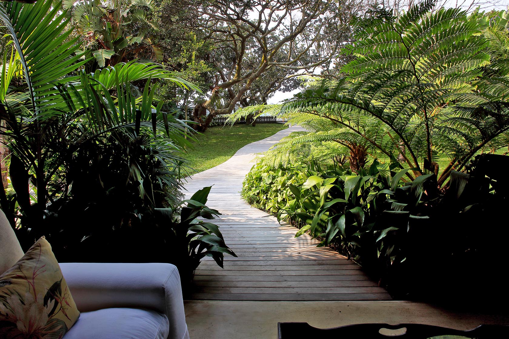 Perfect Hideaways- A peaceful view from a shaded veranda looking onto a curved wooden boardwalk winding through lush tropical gardens with ferns, palms, and manicured lawn cowrie Cottage, Dolphin Coast, Kwazulu- Natal, South Africa