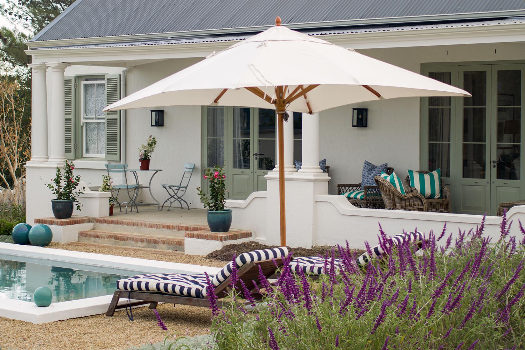 Perfect Hideaways – A charming cottage veranda with soft green shutters and French doors, wicker seating with striped cushions, potted plants, striped sun loungers beside a long pool, and vibrant purple flowers in the foreground. La Cotte Orchard Cottages, Franschhoek South Africa