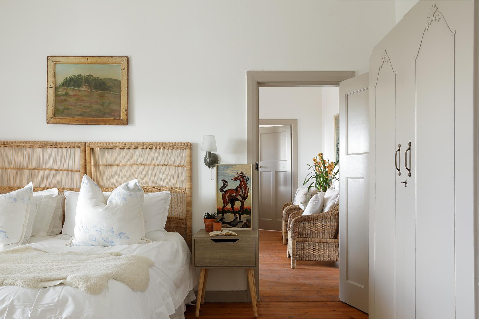 Perfect Hideaways – A peaceful bedroom featuring a woven headboard, airy white linens, and soft blue-patterned pillows. Through the doorway, wicker chairs and greenery add warmth and continuity throughout the cottage. Berit’s Cottage, Churchhaven, South Africa