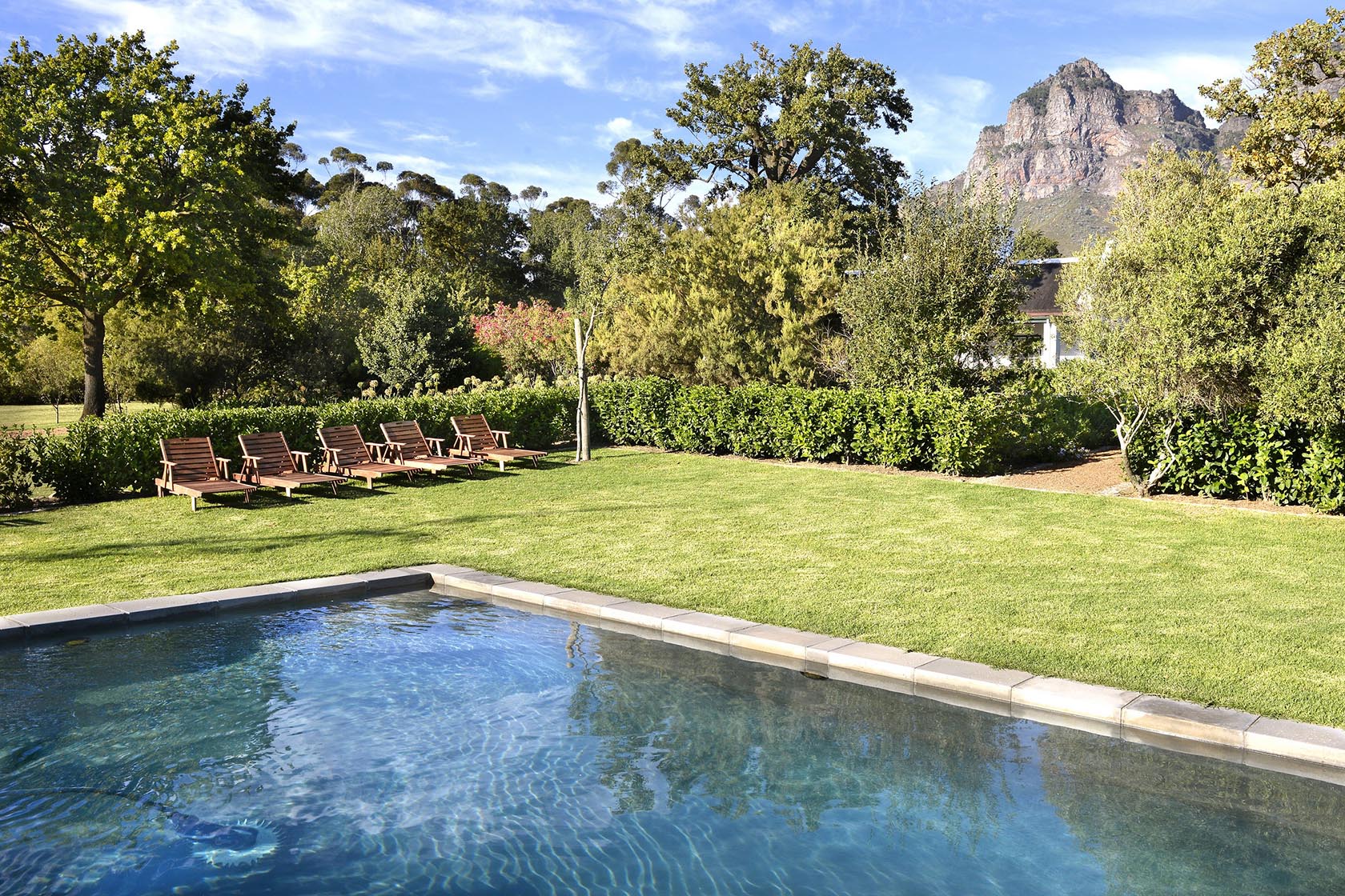 Perfect Hideaways – A serene garden pool reflects the blue sky, bordered by manicured lawn, trees, and a row of wooden sun loungers overlooking distant mountains – Cottage 1685, Franschhoek, Western Cape, South Africa
