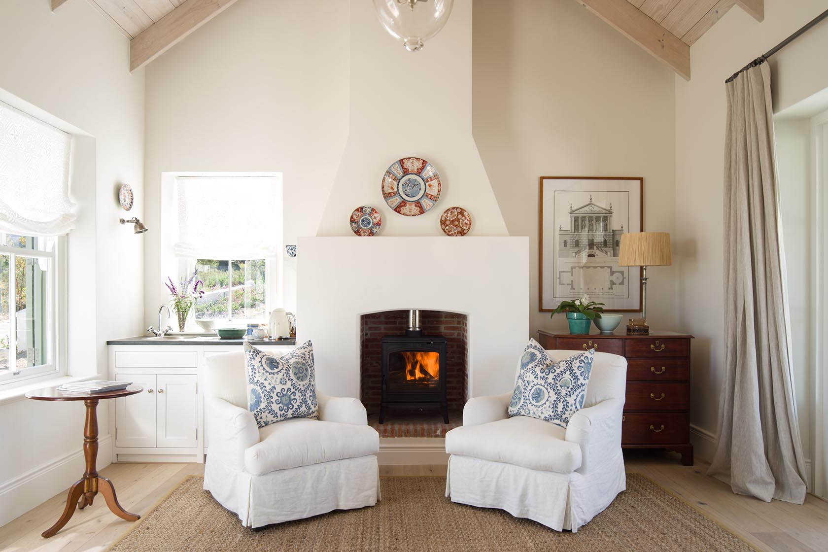 Perfect Hideaways – A cozy living room centered around a wood-burning stove in a white fireplace, flanked by two white armchairs with blue-patterned cushions, warm wooden furniture, framed artwork, and soft natural light creating an inviting atmosphere. La Cotte Orchard Cottages, Franschhoek South Africa