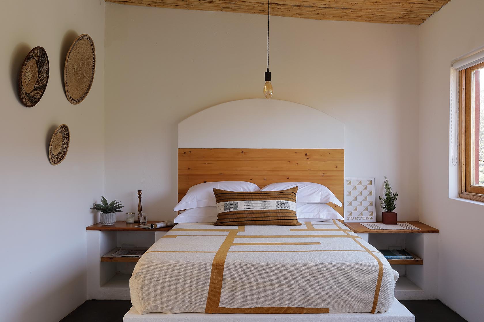 Perfect Hideaways – A serene bedroom with a wooden headboard set against a curved white plaster feature, decorated with woven baskets on the wall. The bed is dressed in a soft, patterned throw and a textured cushion, flanked by simple wooden shelves. Sheperd's Cottage, Prince Albert