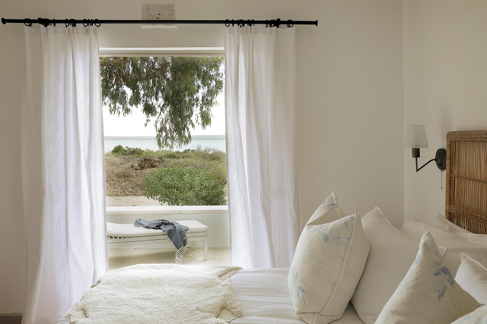 Perfect Hideaways – A bedroom with soft neutral bedding and pillows, opening onto an outdoor scene through flowing white curtains. A simple bench sits outside beneath a eucalyptus tree, with views of the lagoon shimmering in the distance. Berit’s Cottage, Churchhaven, South Africa.