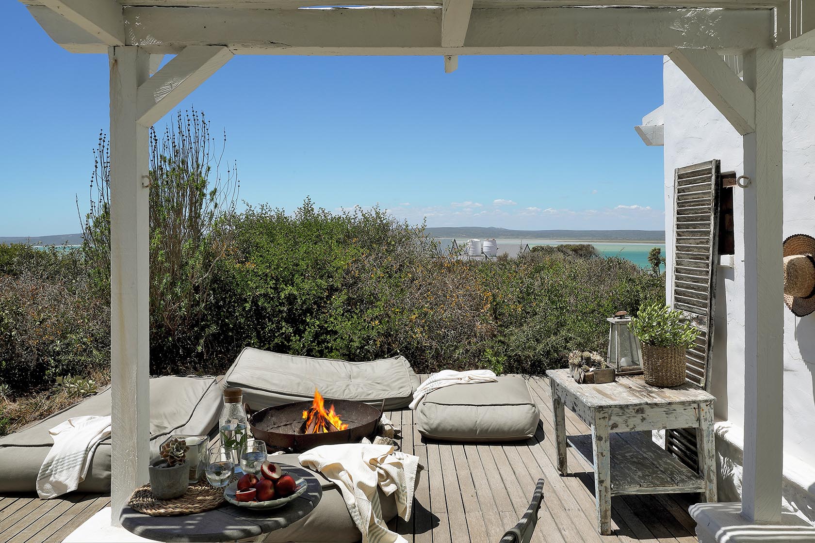 Perfect Hideaways–Deck lounge with cushioned sunbeds, firepit, rustic table, and views over fynbos toward the lagoon. Church Cottage, Churchhaven, South Africa