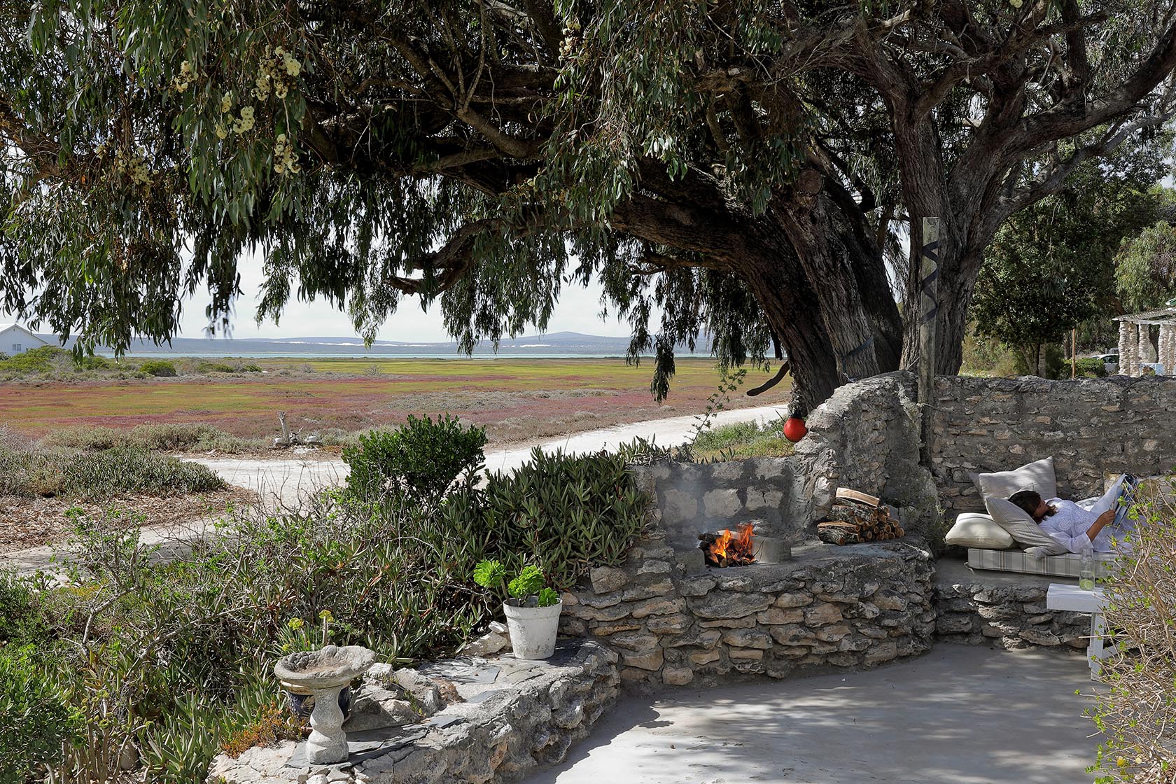 Perfect Hideaways. A stone-walled patio shaded by a large leafy tree, featuring a built-in stone braai fireplace with a crackling fire. A person relaxes on an adjacent cushioned stone bench with a book, looking out over a vast coastal landscape and lagoon, Berit's Cottage, Churchhaven