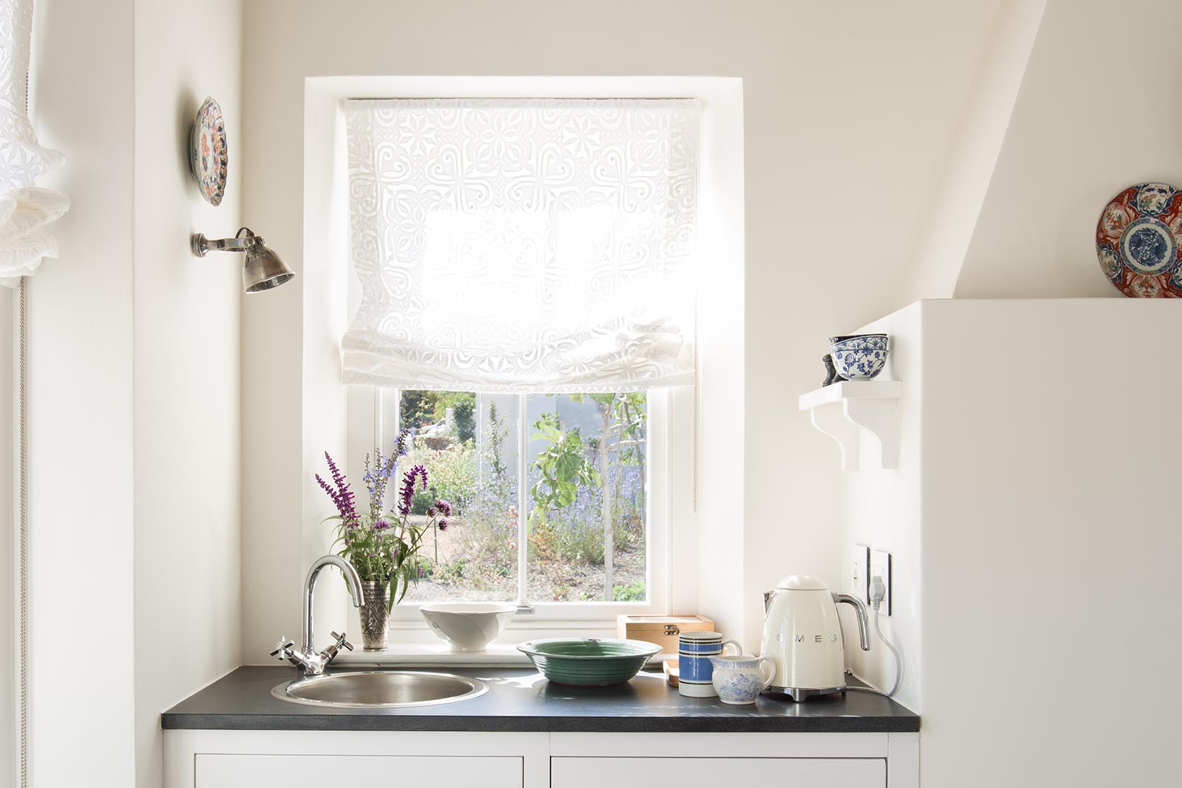 Perfect Hideaways – A bright kitchenette area with a black countertop, white cabinetry, a window covered with a delicate white Roman blind, and ceramics and flowers arranged neatly beside a cream-colored SMEG kettle, all bathed in soft daylight. La Cotte Orchard Cottages, Franschhoek South Africa