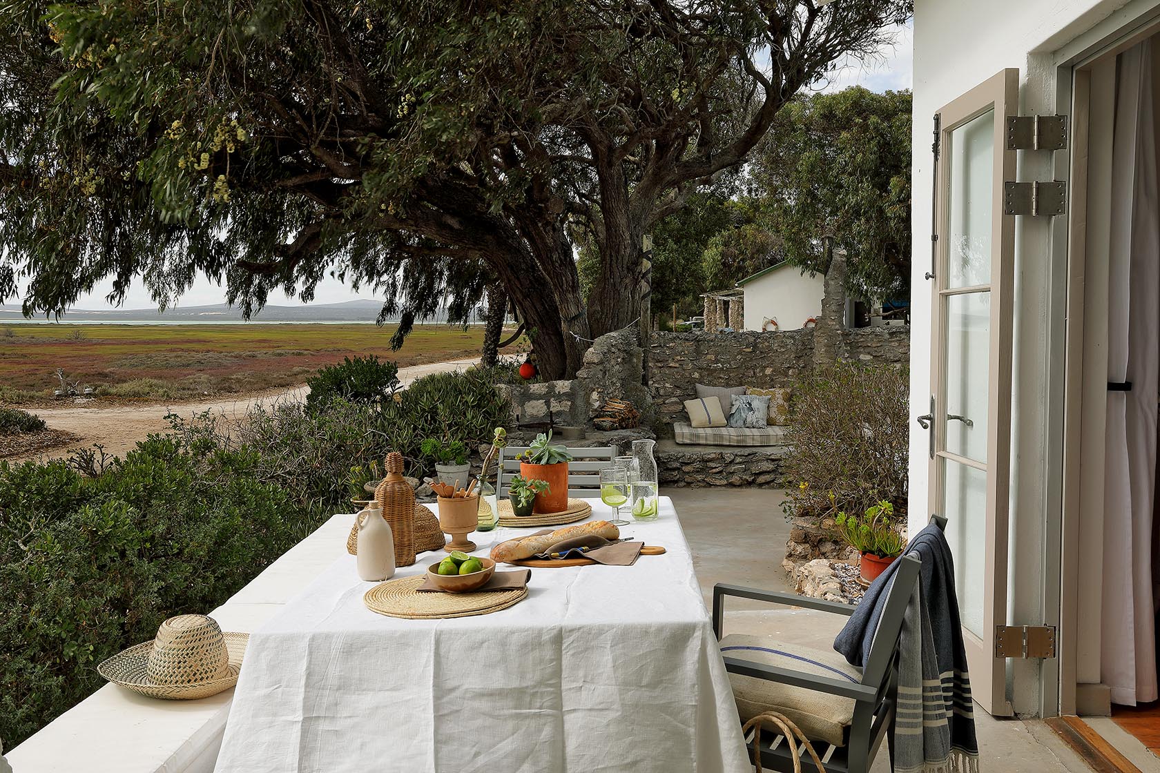Perfect Hideaways – A natural, rustic outdoor dining setup beneath the shade of a large tree, with a long white-draped table dressed in woven mats, pottery, bread, fruit, and potted succulents. Beyond the patio, the marshland and lagoon stretch toward distant hills. Berit’s Cottage, Churchhaven, South Africa.