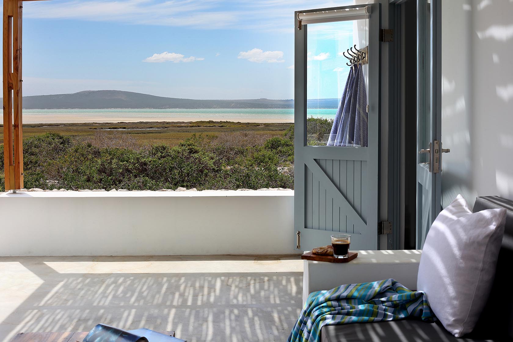 Perfect Hideaways – A shaded outdoor seating area with soft light filtering through a pergola, looking out toward a serene lagoon and distant hills. A blue half-stable door stands open with towels hanging inside, while a cushioned bench with a white pillow and striped throw holds a tray with coffee; Johanns Cottage, Churchhaven.