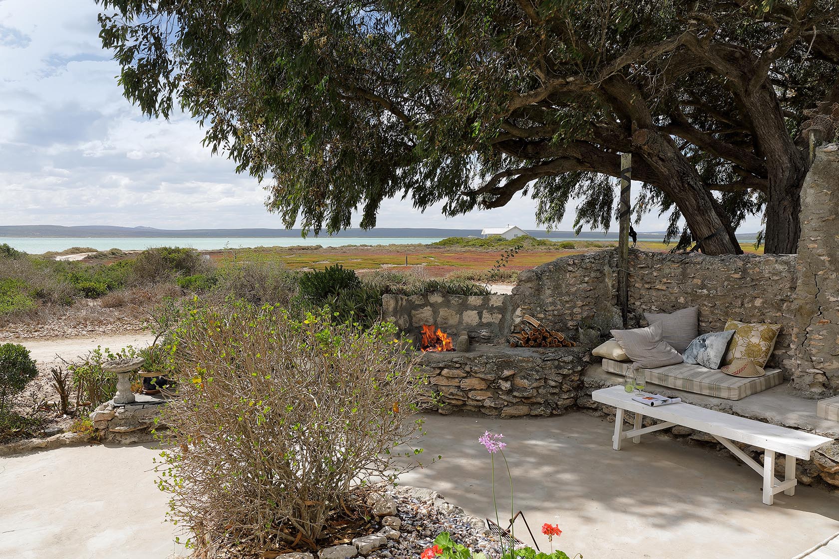 Perfect Hideaways. A stone-paved outdoor terrace under the shade of a large tree, showcasing a stone fireplace with a burning fire, a built-in seating area with pillows, a white wooden bench, and an expansive view of the surrounding salt marshes and lagoon, Berit's Cottage, Churchhaven