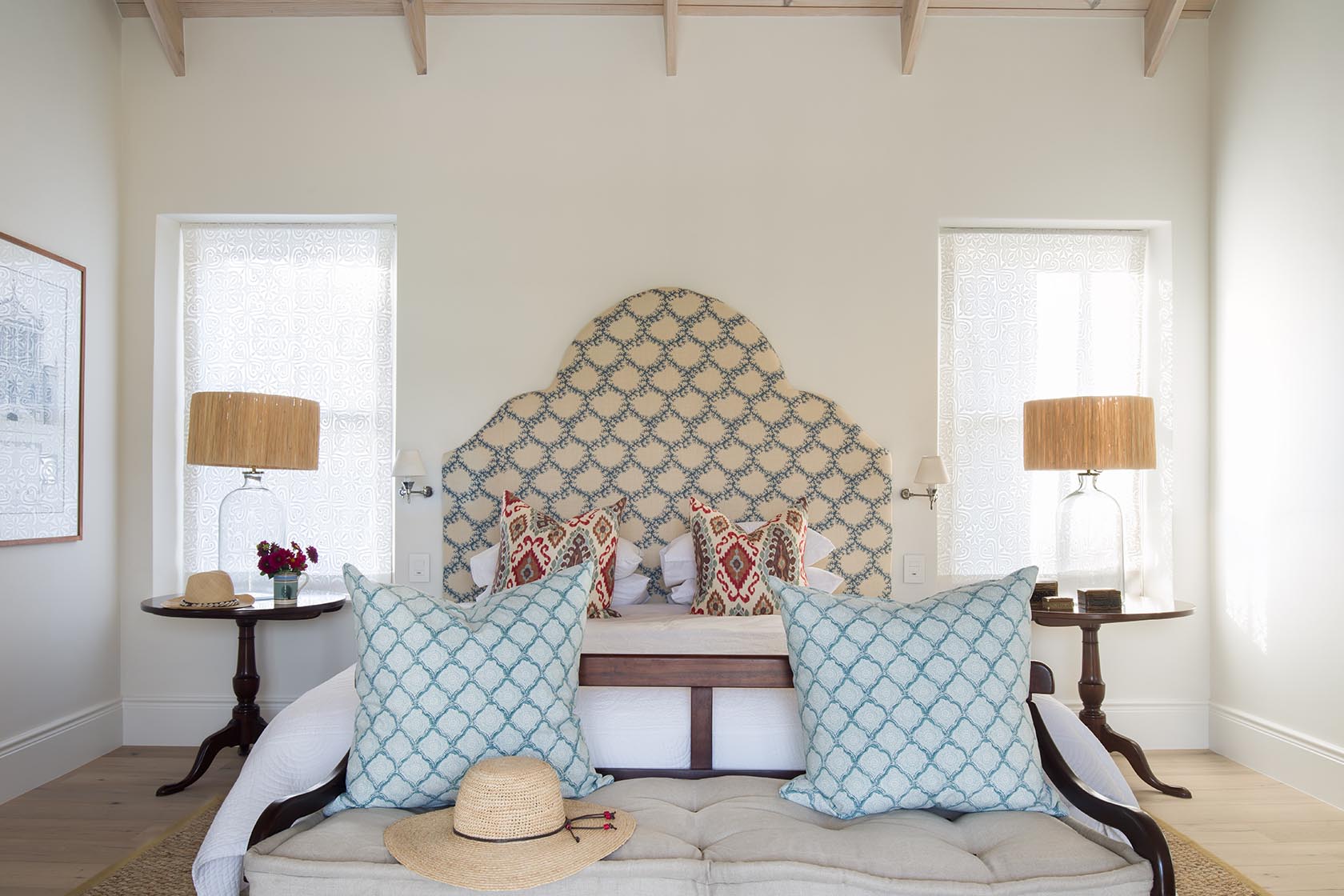 Perfect Hideaways – A serene bedroom with a scalloped upholstered headboard in soft blue and cream, flanked by two tall windows draped with delicate white patterned blinds. The bed is layered with textured pillows and crisp white linens, with a cushioned bench at the foot holding a straw sun hat. Wooden side tables with glass lamps and fresh flowers complete the airy, inviting look. La Cotte Orchard Cottages, Franschhoek South Africa
