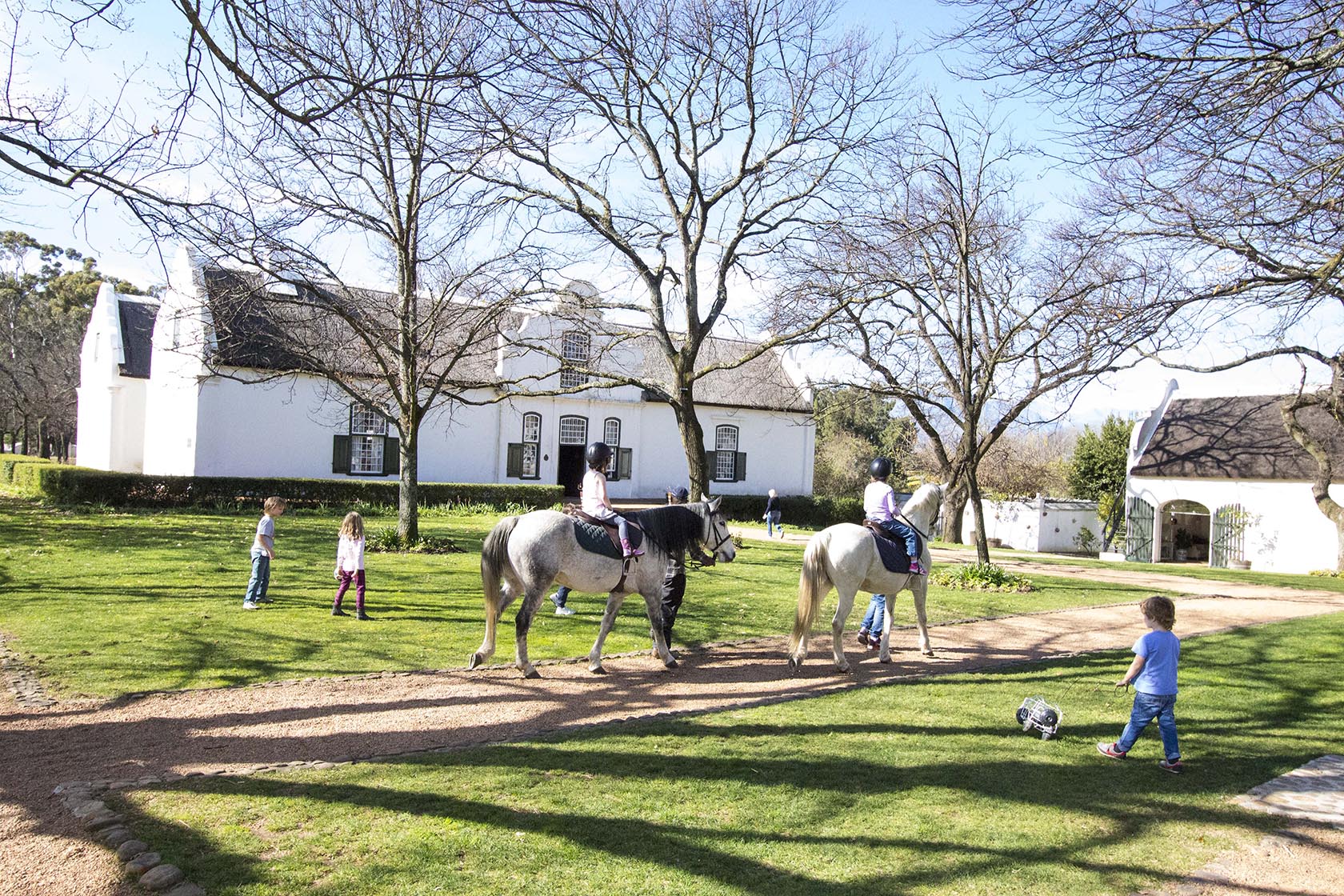 Perfect Hideaways – Children play on a sunny lawn while two young riders on ponies pass in front of a white Cape Dutch homestead framed by bare winter trees – Cottage 1685, Franschhoek, Western Cape, South Africa