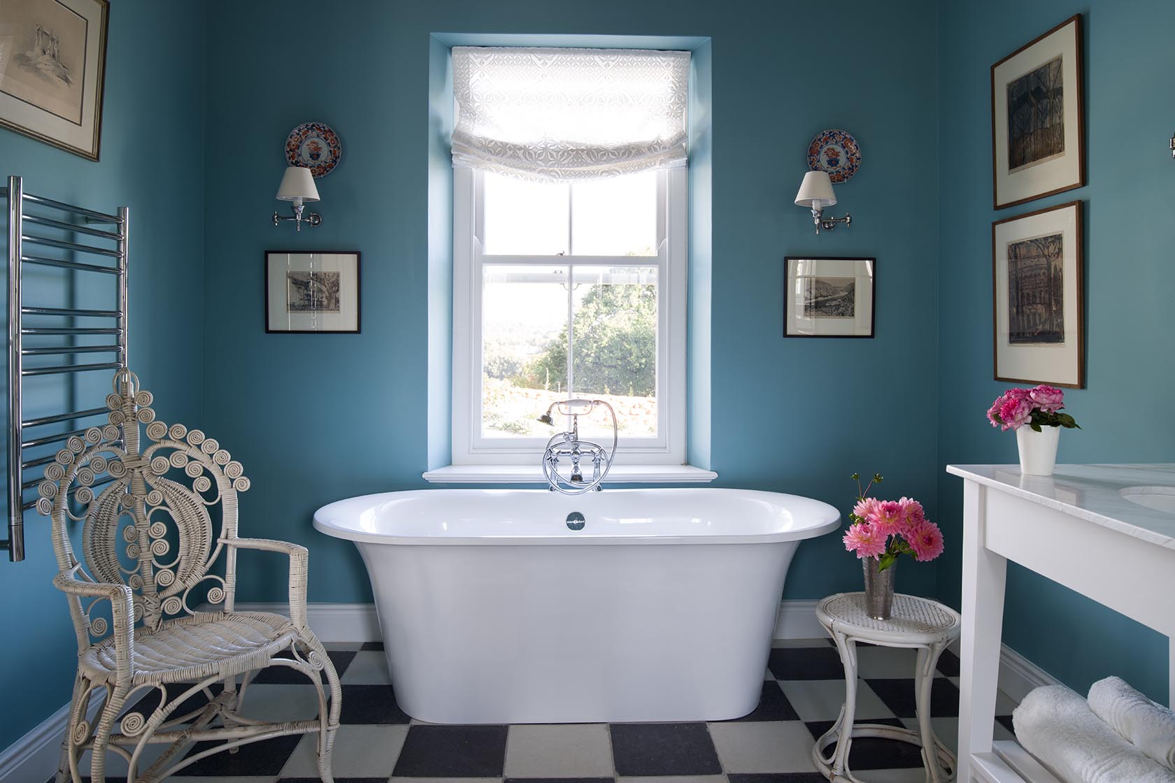 Perfect Hideaways – A bold blue bathroom featuring a freestanding white tub beneath a bright window, surrounded by framed artwork. A decorative wicker chair, checkerboard floor tiles, and fresh pink flowers add charm to the space, blending vintage style with crisp elegance. La Cotte Orchard Cottages, Franschhoek South Africa