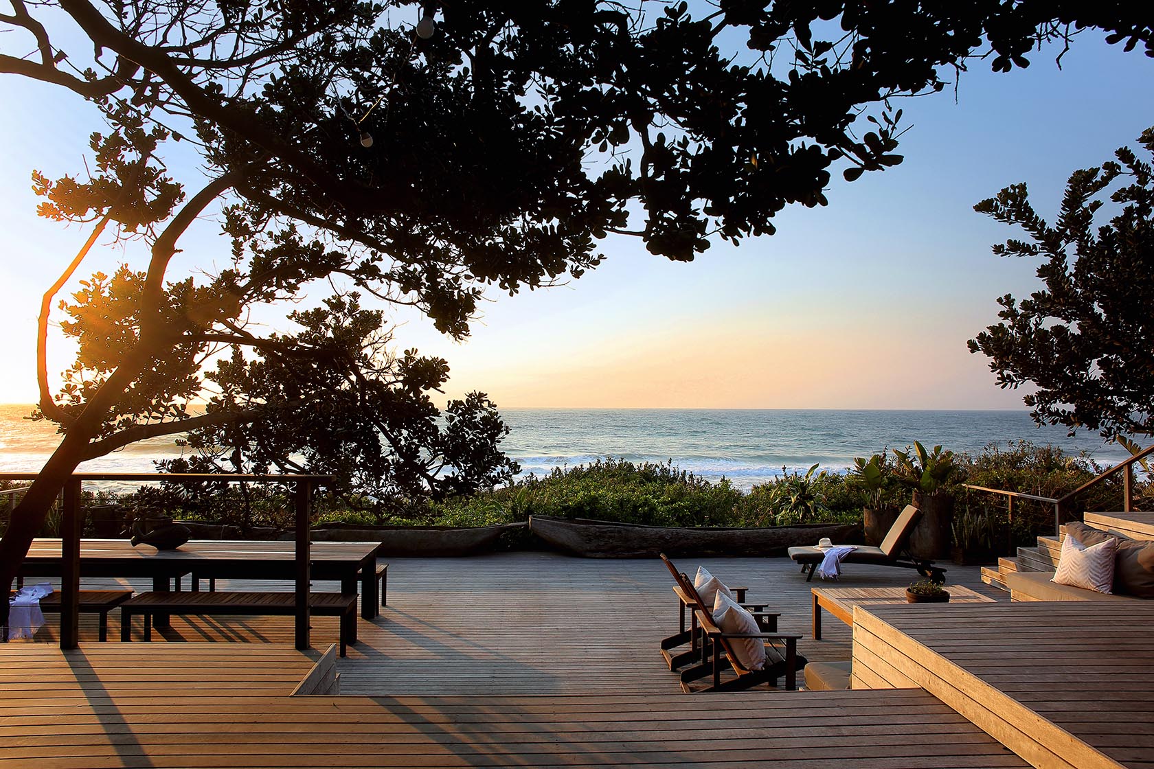 Perfect Hideaways- A spacious ocean-facing wooden deck at sunset with outdoor seating, dining tables, and loungers framed by coastal trees and warm golden light cowrie Cottage, Dolphin Coast, Kwazulu- Natal, South Africa