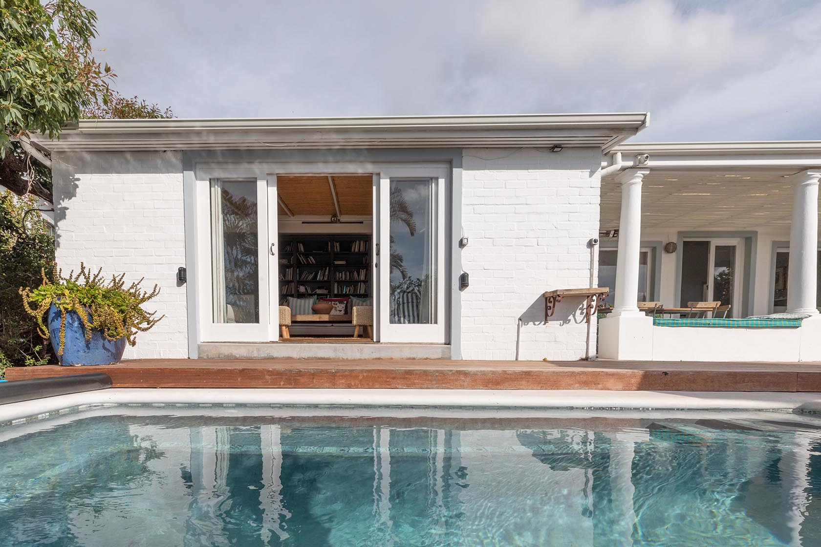 Perfect Hideaways – Poolside view of a white cottage with large sliding glass doors opening from a library room, framed by greenery and reflected in the clear turquoise pool water below. Boulders Cottage, Simons Town