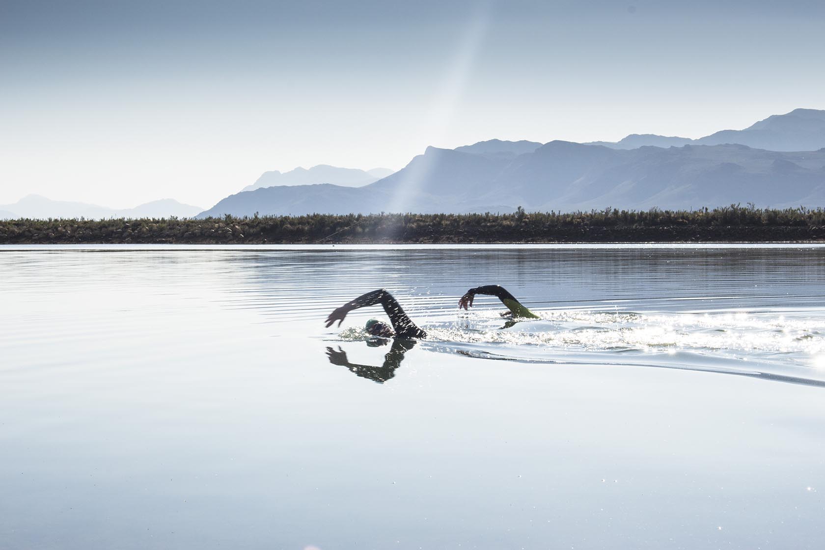 Perfect Hideaways – Two swimmers in wetsuits glide across a still lake under bright morning light, with layered mountain silhouettes stretching across the horizon – Cottage 1685, Franschhoek, Western Cape, South Africa