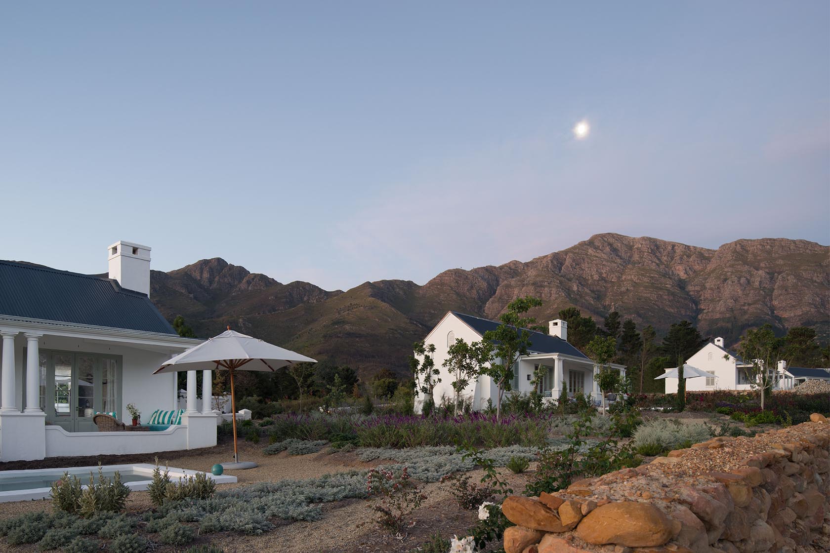 Perfect Hideaways – A peaceful twilight view of multiple white cottages nestled among lush gardens, with an umbrella-shaded terrace and pool in the foreground. Majestic mountains rise in the distance under a soft evening sky with the faint glow of the moon. La Cotte Orchard Cottages, Franschhoek South Africa