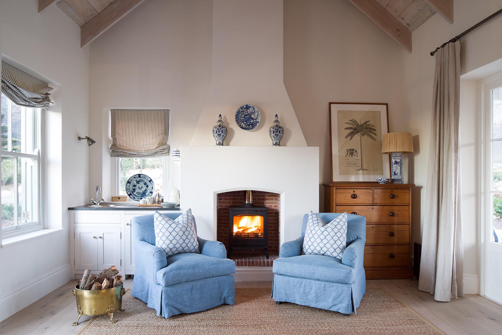 Perfect Hideaways – A cozy sitting room with two soft blue armchairs facing a wood-burning stove set within a white fireplace. Striped Roman blinds, blue-and-white ceramic pieces, and warm wooden furniture give the room a refined country charm. La Cotte Orchard Cottages, Franschhoek South Africa