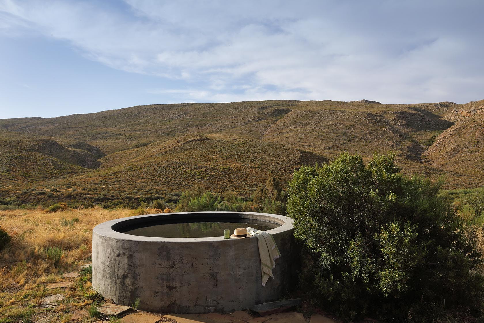Perfect Hideaways – A round concrete plunge pool sits at the edge of the landscape, its still water reflecting the sky. A sun hat and towel rest on the edge, surrounded by natural scrub and rolling Karoo hills. Sheperd's Cottage, Prince Albert