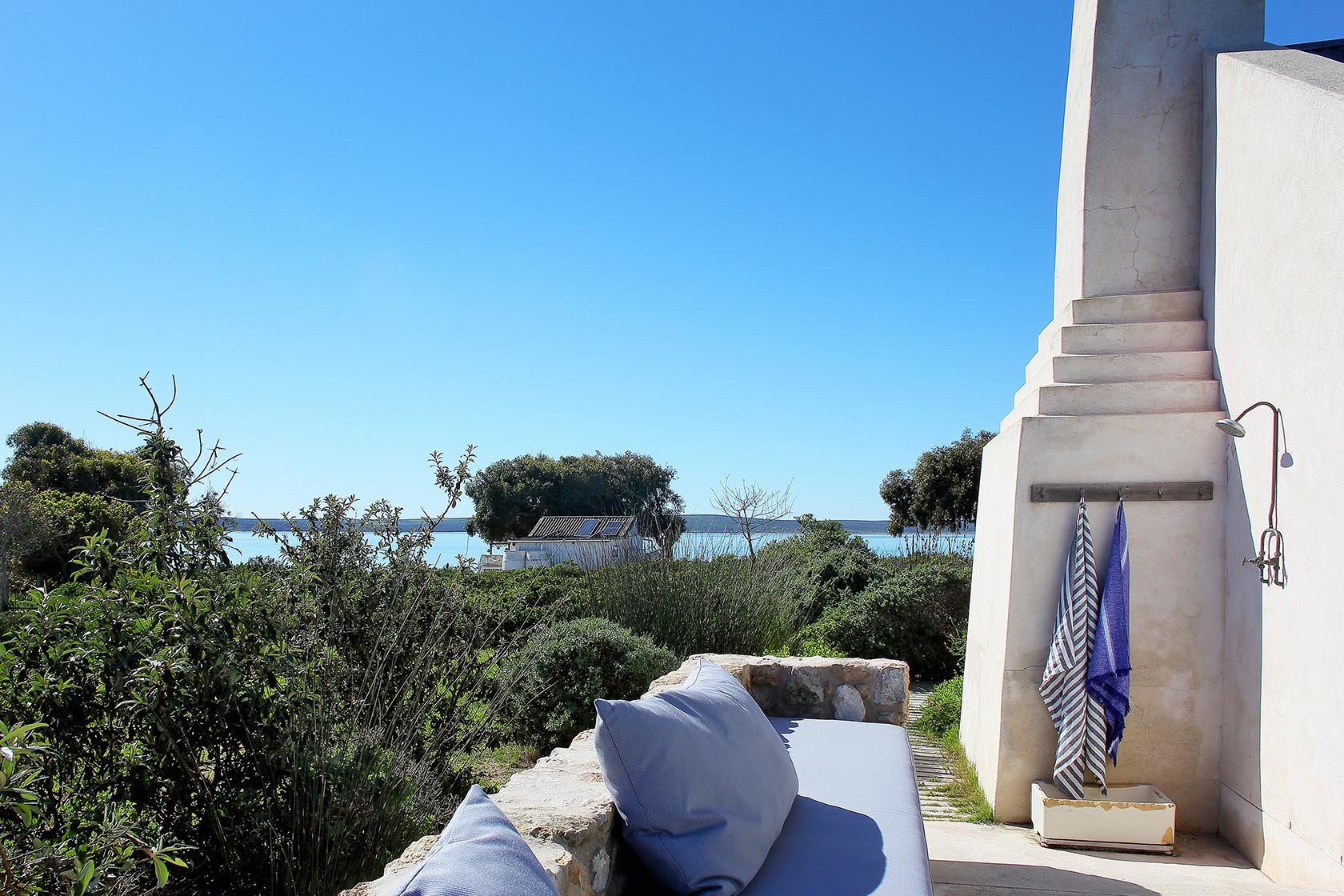 Perfect Hideaways- An outdoor shower nook beside the cottage chimney, with striped towels hanging on hooks, cushioned stone seating, and expansive views over fynbos toward the lagoon. Milkwood Cottage, Churchhaven, South Africa