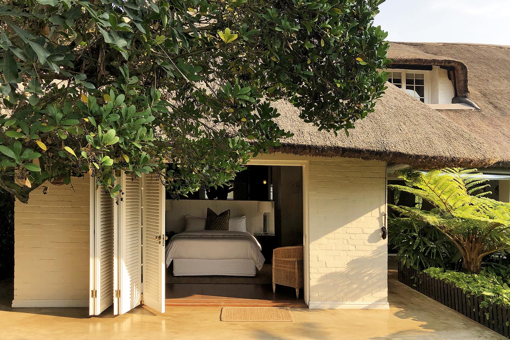 Perfect Hideaways- A thatched-roof cottage bedroom with open doors revealing a neatly dressed bed and surrounding lush garden greenery cowrie Cottage, Dolphin Coast, Kwazulu- Natal, South Africa