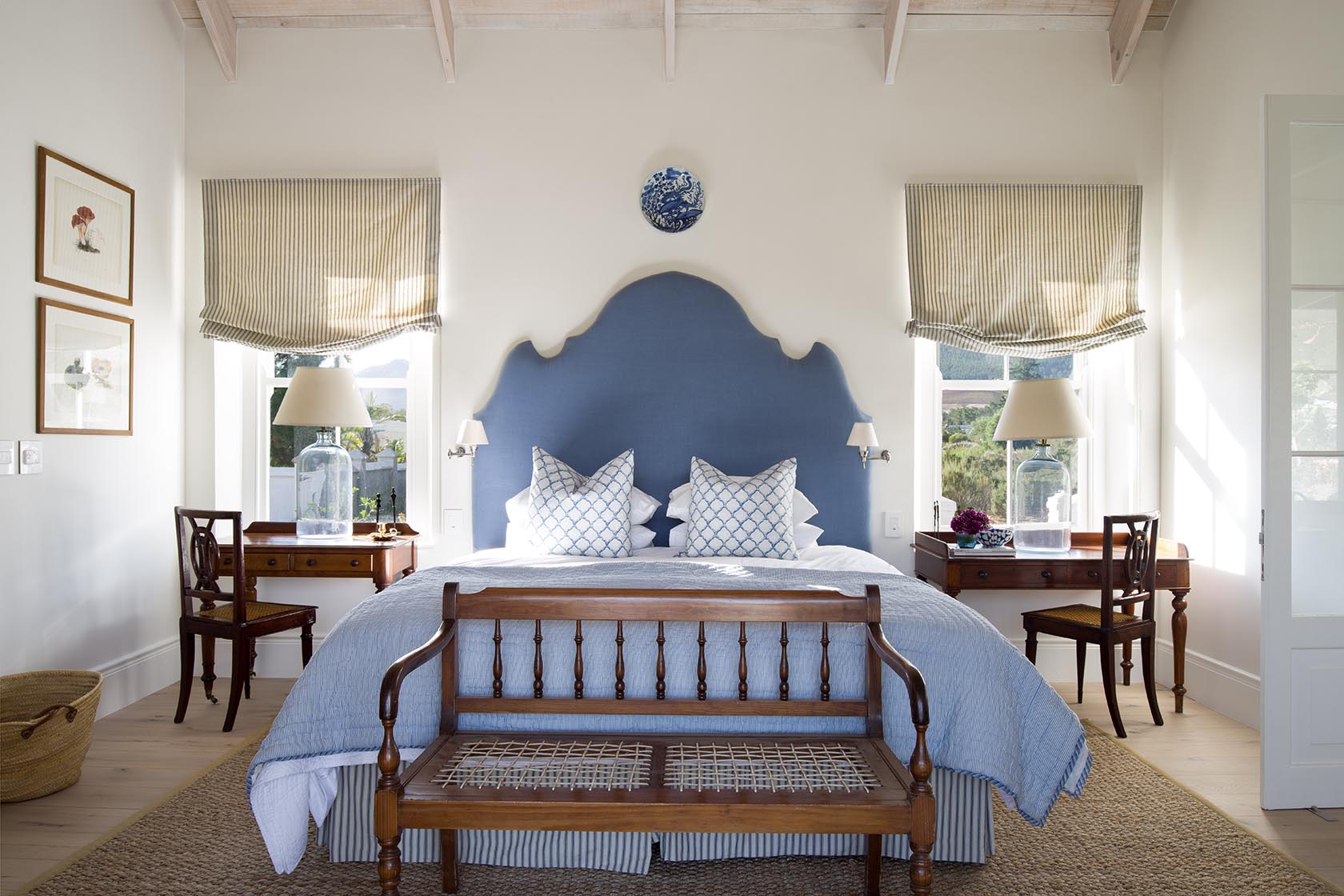 Perfect Hideaways – A bright bedroom with a striking deep blue scalloped headboard, twin desks flanking the bed, striped Roman blinds, and blue-accented bedding. A wooden bench sits at the foot of the bed, creating a balanced, elegant composition. La Cotte Orchard Cottages, Franschhoek South Africa