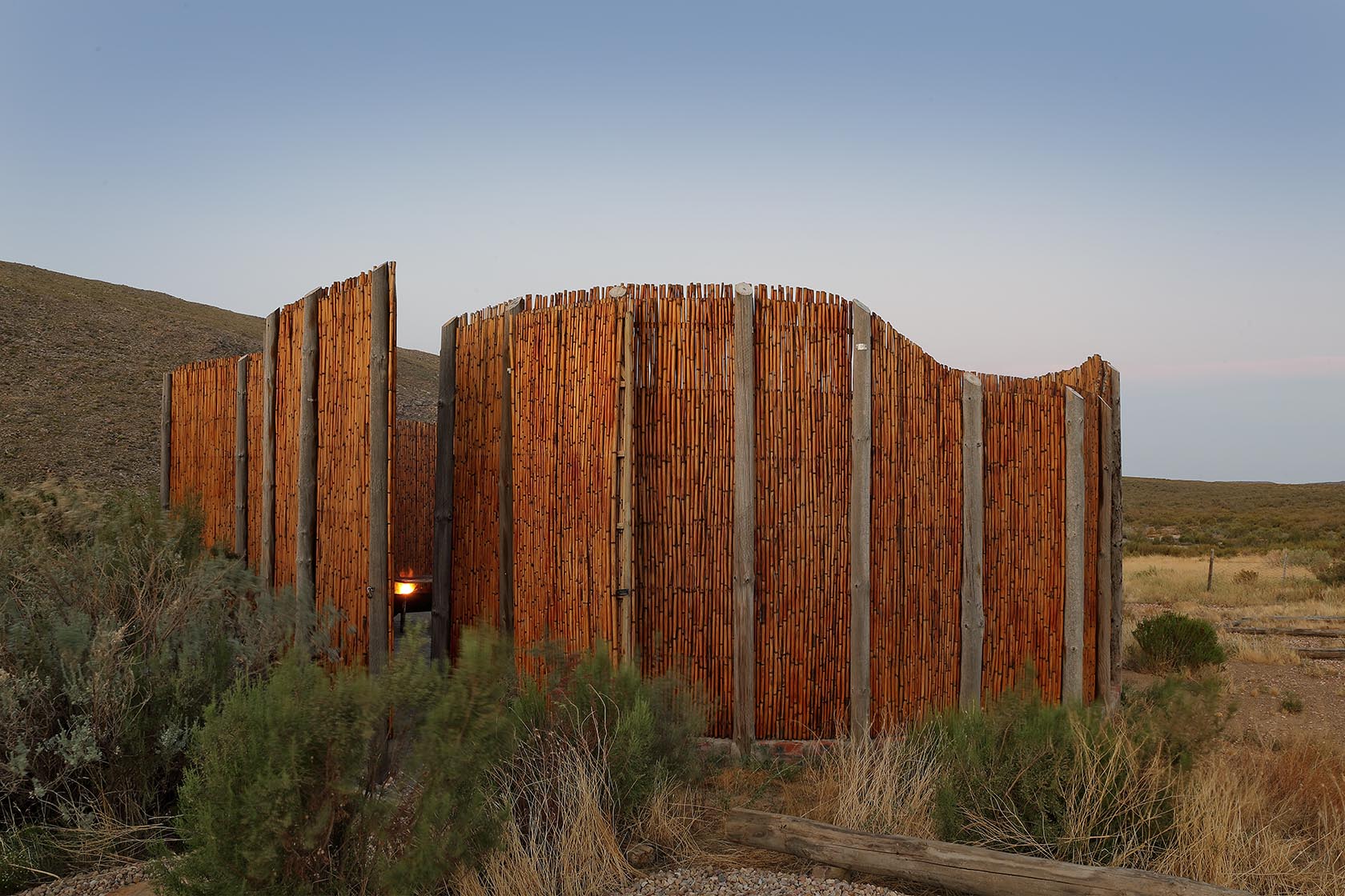 Perfect Hideaways – A tall, sculptural enclosure made of vertical timber slats stands in the open veld, its organic form blending with the muted colours of the Karoo. Soft twilight settles over the scene. Sheperd's Cottage, Prince Albert