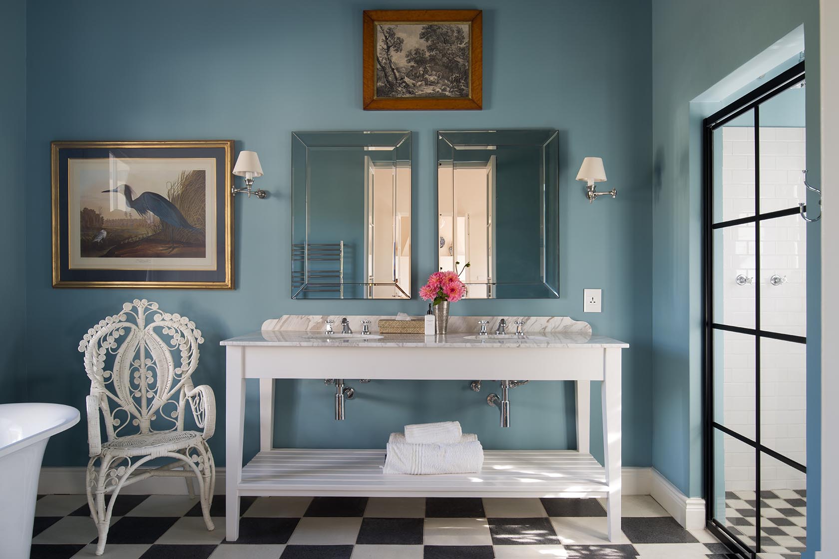 Perfect Hideaways – A sophisticated blue-toned bathroom featuring a double vanity with marble countertop, twin mirrored wall panels, a decorative wicker chair, and checkered floor tiles. Artwork and fresh pink flowers add color and softness to the refined design. La Cotte Orchard Cottages, Franschhoek South Africa
