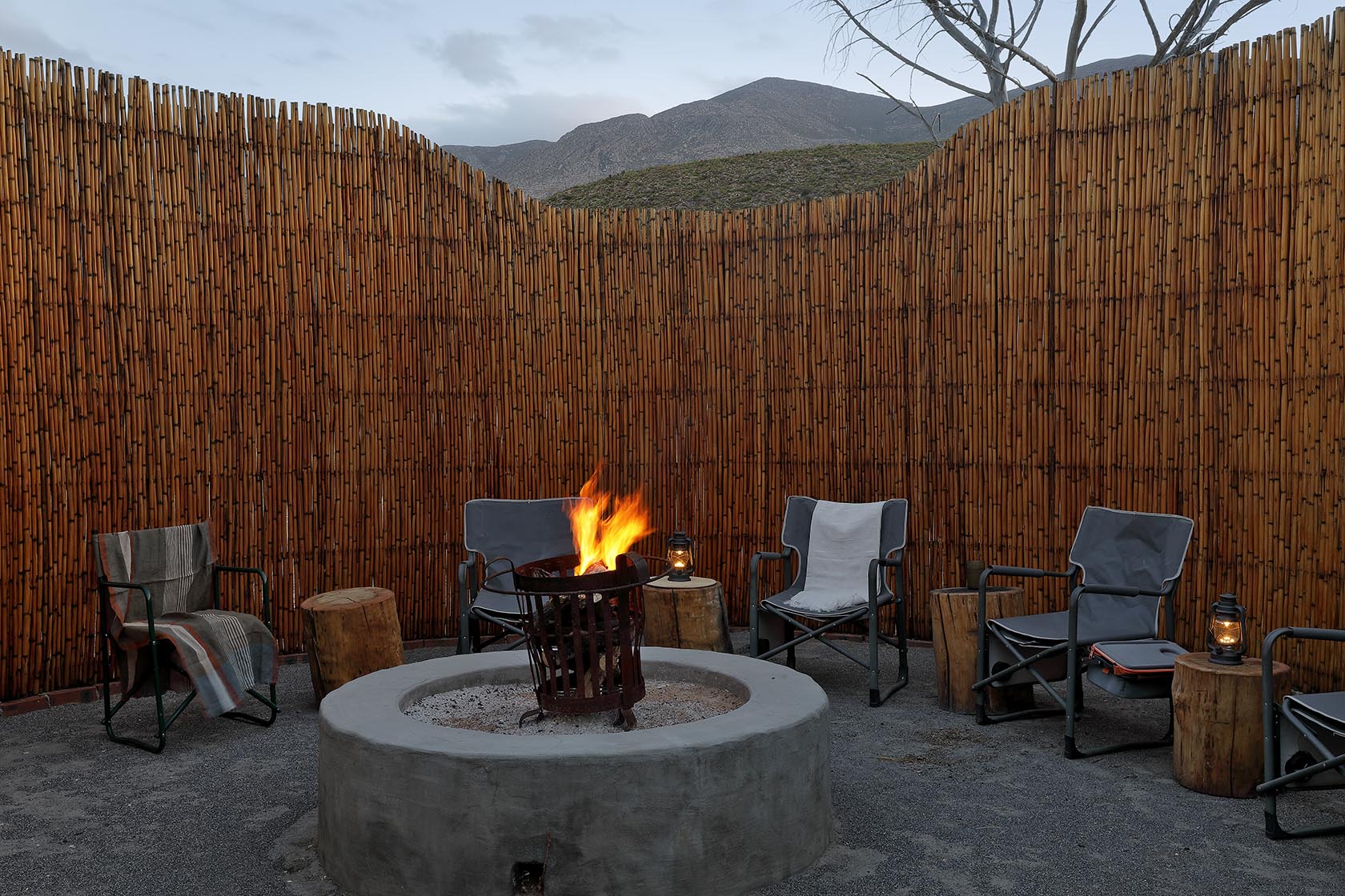 Perfect Hideaways – An intimate circular fire pit area enclosed by tall timber slats, with canvas chairs arranged around a glowing fire. Lanterns provide warm light against the backdrop of rugged hills. Sheperd's Cottage, Prince Albert