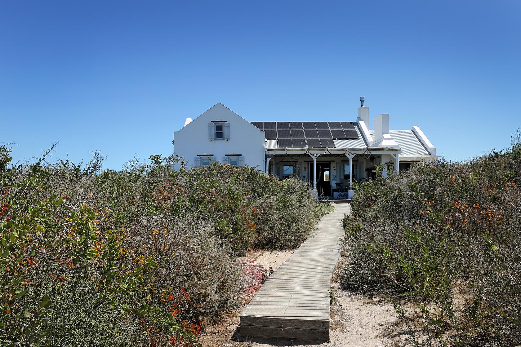 Perfect Hideaways–View of the whitewashed cottage from a boardwalk cutting through coastal fynbos under a bright blue sky. Church Cottage, Churchhaven, South Africa