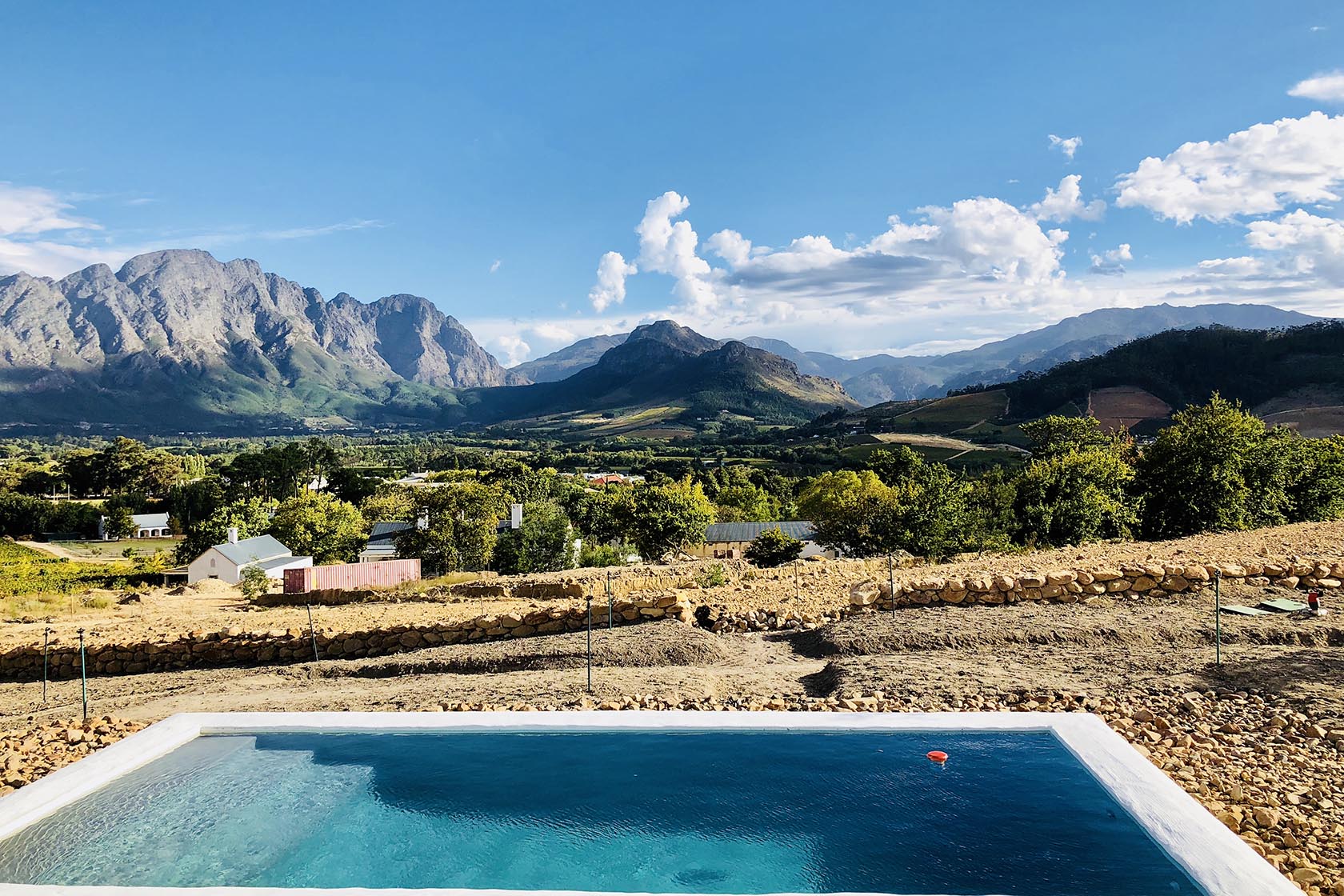 Perfect Hideaways – A dramatic mountain landscape stretching across the horizon, viewed from a crystal-clear turquoise pool in the foreground. Rolling vineyards, stone terraces, and scattered farm buildings nestle into the scenic valley under a bright blue sky. La Cotte Orchard Cottages, Franschhoek South Africa