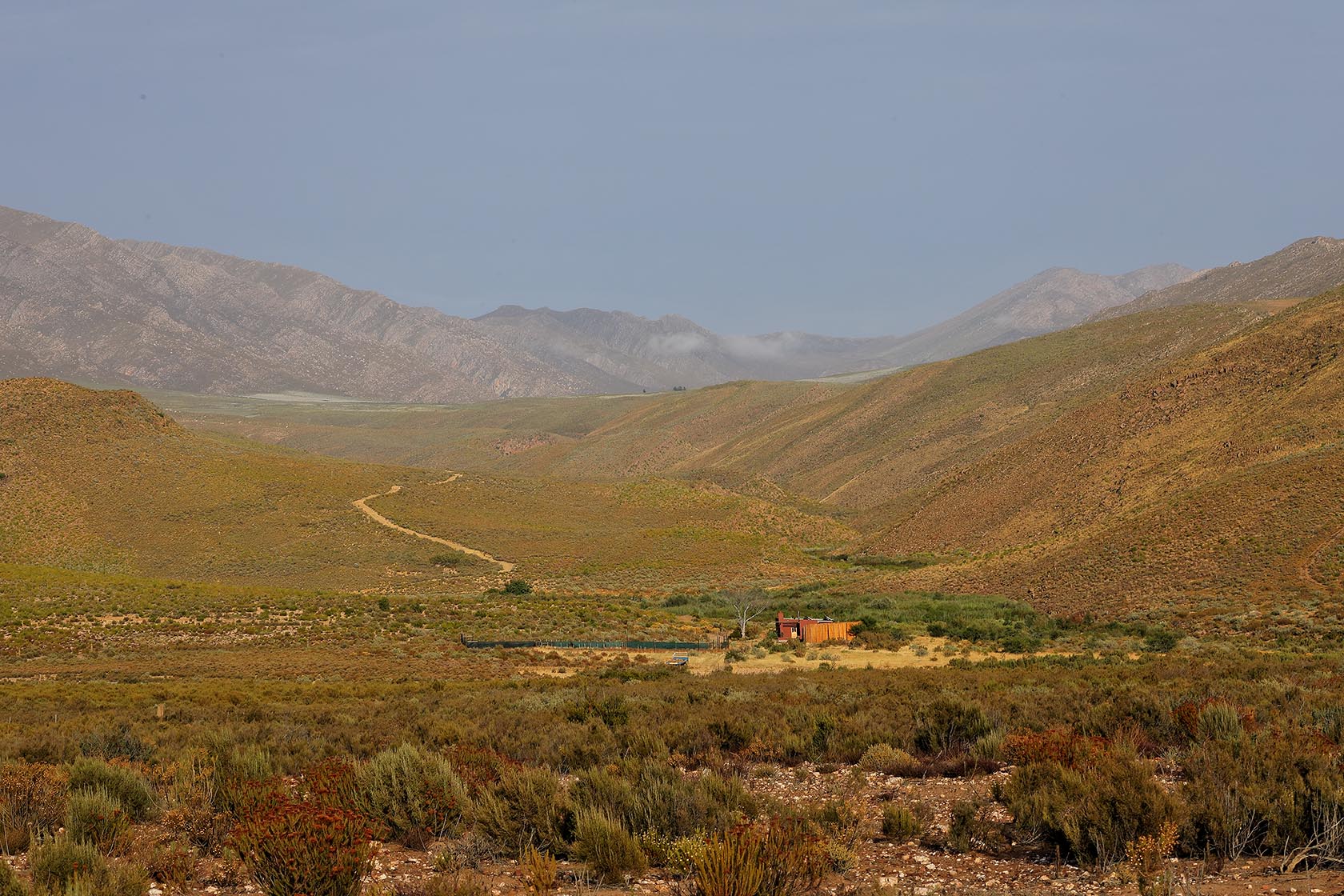 Perfect Hideaways – A wide valley view shows the cottage nestled among scrubland at the base of sweeping, ochre-coloured mountains. A dusty track winds through the landscape beneath a soft blue sky. Sheperd's Cottage, Prince Albert