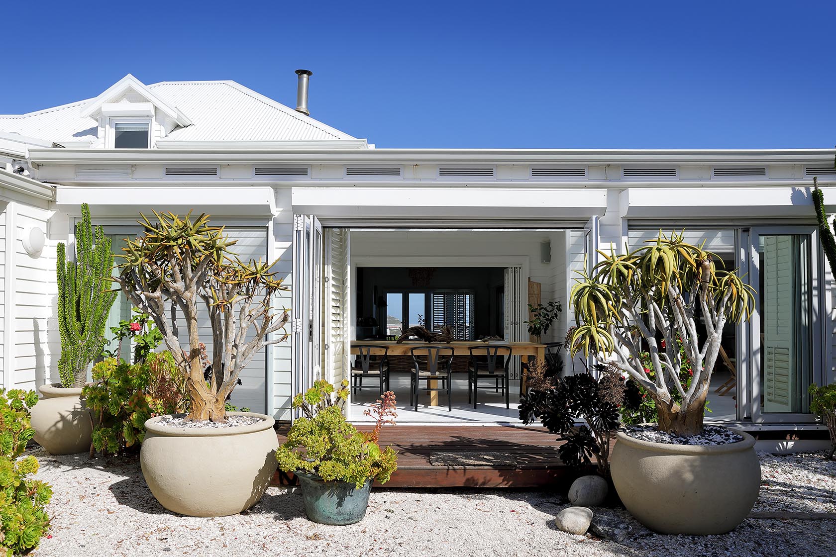 Perfect Hideaways - Exterior view of the white coastal cottage with open doors revealing the dining area, framed by sculptural succulents, gravel pathways and blue skies Cape Cottage_Yzerfontein_West Coast_Western Cape_South Africa
