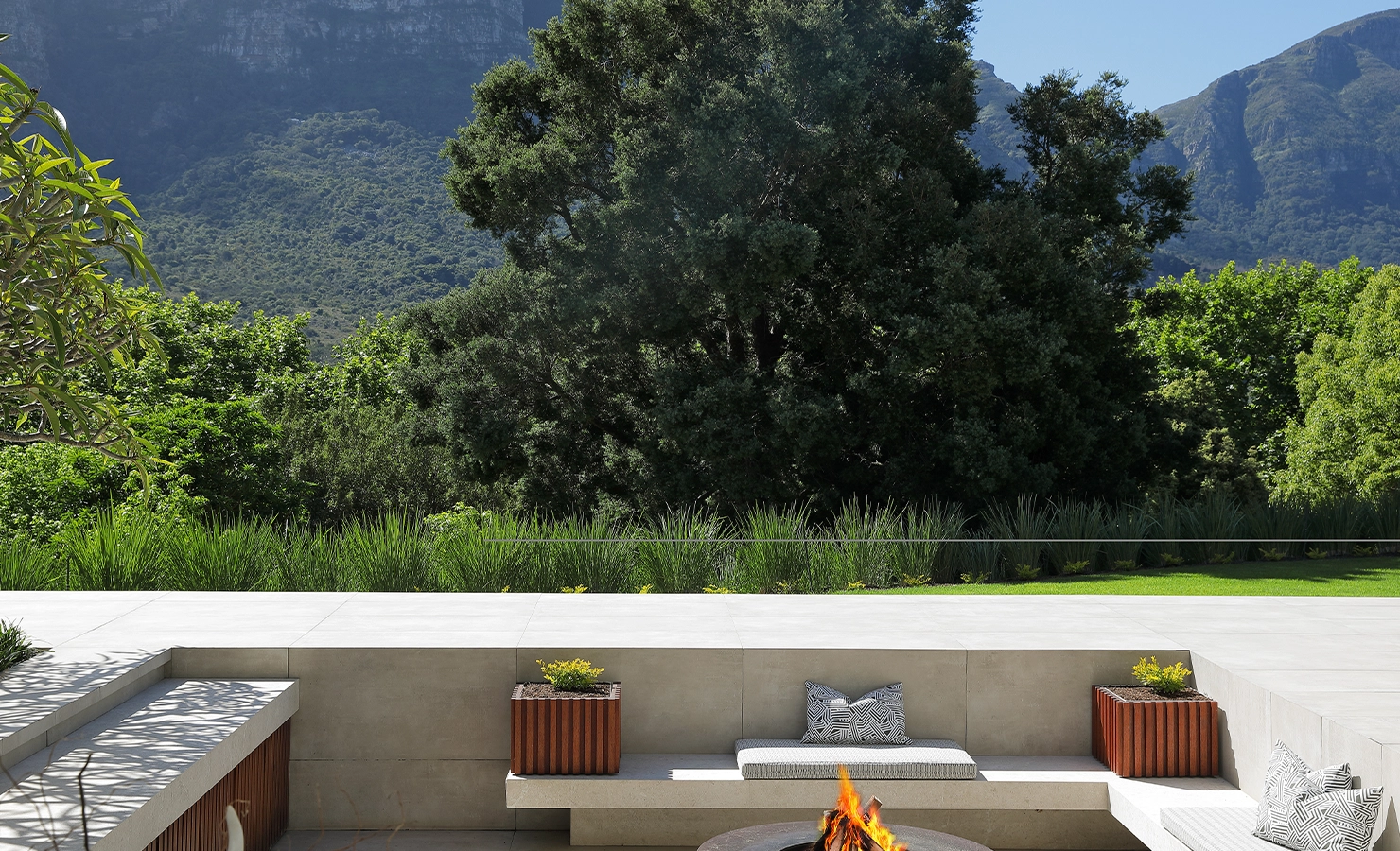 Perfect Hideaways A tranquil outdoor firepit area with built-in stone seating, patterned cushions, and timber planter boxes, overlooking dense green trees and a dramatic mountain backdrop Pure Living, Bishopscourt