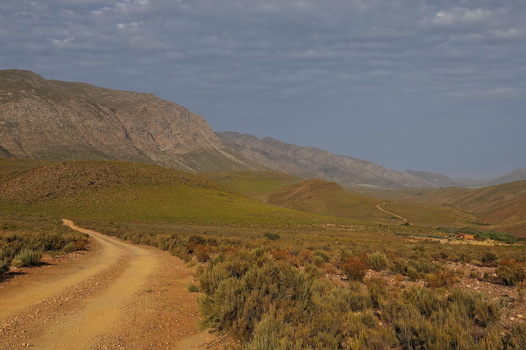 Perfect Hideaways – A long, winding gravel road leads through the expansive Karoo landscape toward distant rocky mountains. Low shrubs and warm-toned vegetation stretch across the valley floor. Sheperd's Cottage, Prince Albert