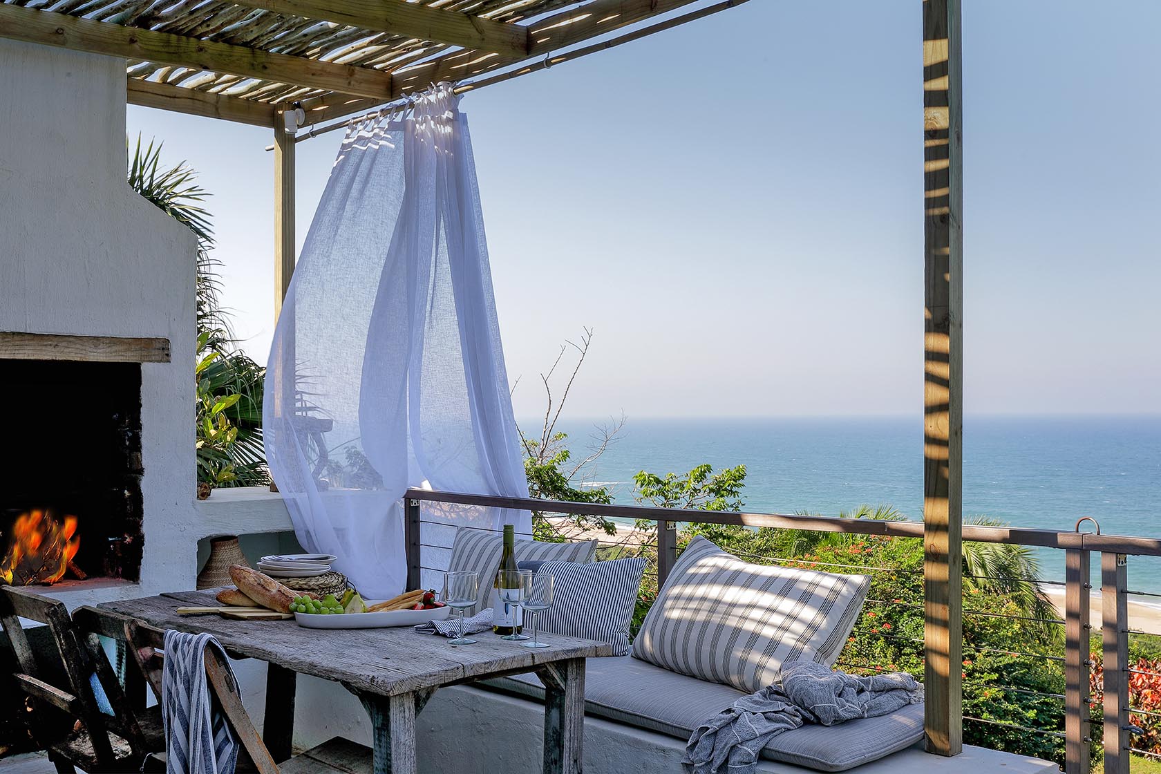 Perfect Hideaways – An inviting outdoor dining terrace overlooking the ocean, featuring a rustic wooden table set with fresh food and wine beside a built-in fireplace, flowing white curtains, and cushioned seating under a slatted pergola – Cottage by the Sea, Elysium.