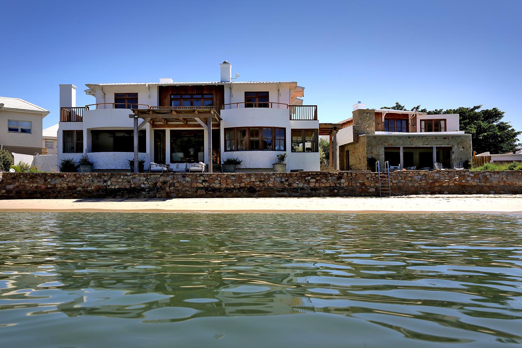 Perfect Hideaways: Lagoon-front exterior showing curved balconies, pergola deck and stone sea wall above the sand, Knysna, Tides Reach
