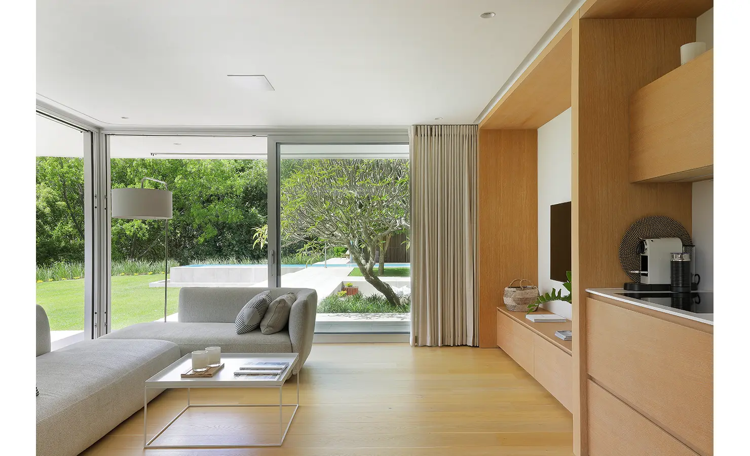 Perfect Hideaways A minimalist living room with pale wood finishes, soft grey seating, and expansive glass doors opening to a terrace, showcasing a sculptural tree and manicured lawn outside Pure Living, Bishopscourt