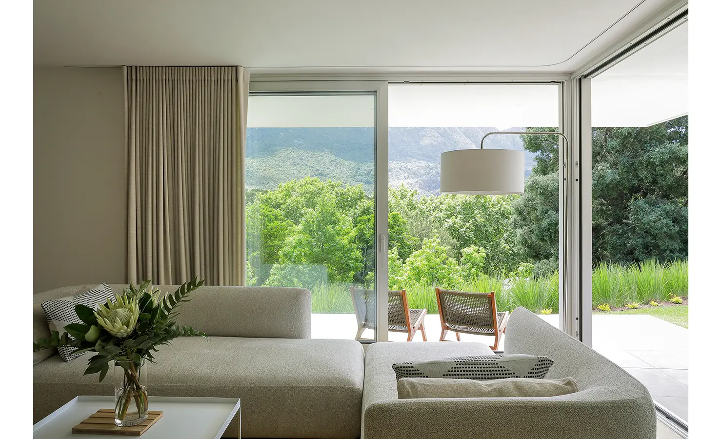 Perfect Hideaways A cozy living room with plush pale sectional sofas, soft patterned cushions, and expansive sliding doors revealing mountain views and lush trees Pure Living, Bishopscourt