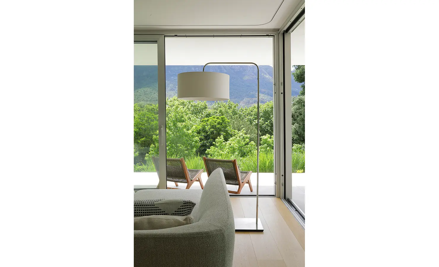Perfect Hideaways A serene view through large sliding glass doors reveals two woven loungers on a sunlit terrace, surrounded by lush greenery and framed by distant mountains Pure Living, Bishopscourt