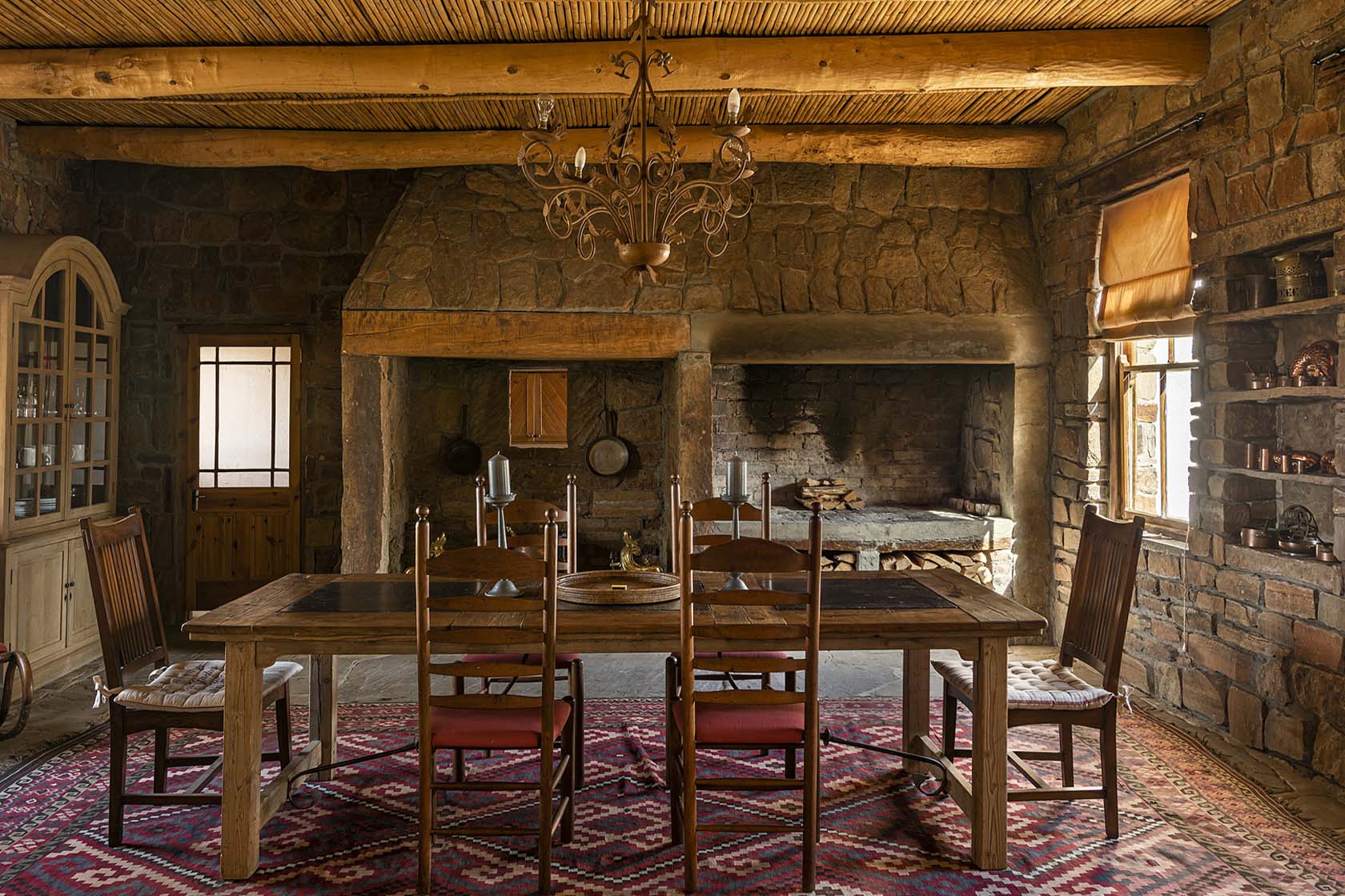 Perfect Hideaways- A dramatic stone-walled dining room with a large wooden table, high-backed chairs, an ornate chandelier, a woven rug, and a cavernous built-in hearth that evokes historic Karoo farm architecture. Karoo Cottage, Sneeuberg Nature Reserve, Eastern Cape, South Africa