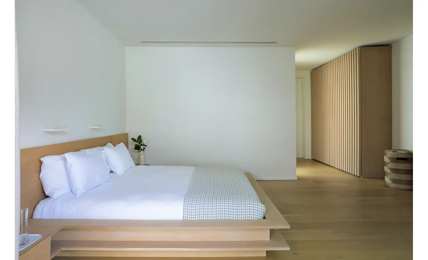 Perfect Hideaways A serene minimalist bedroom with a low timber platform bed dressed in crisp white linens, soft natural light, and a corridor lined with pale wooden slatted cabinetry Pure Living, Bishopscourt