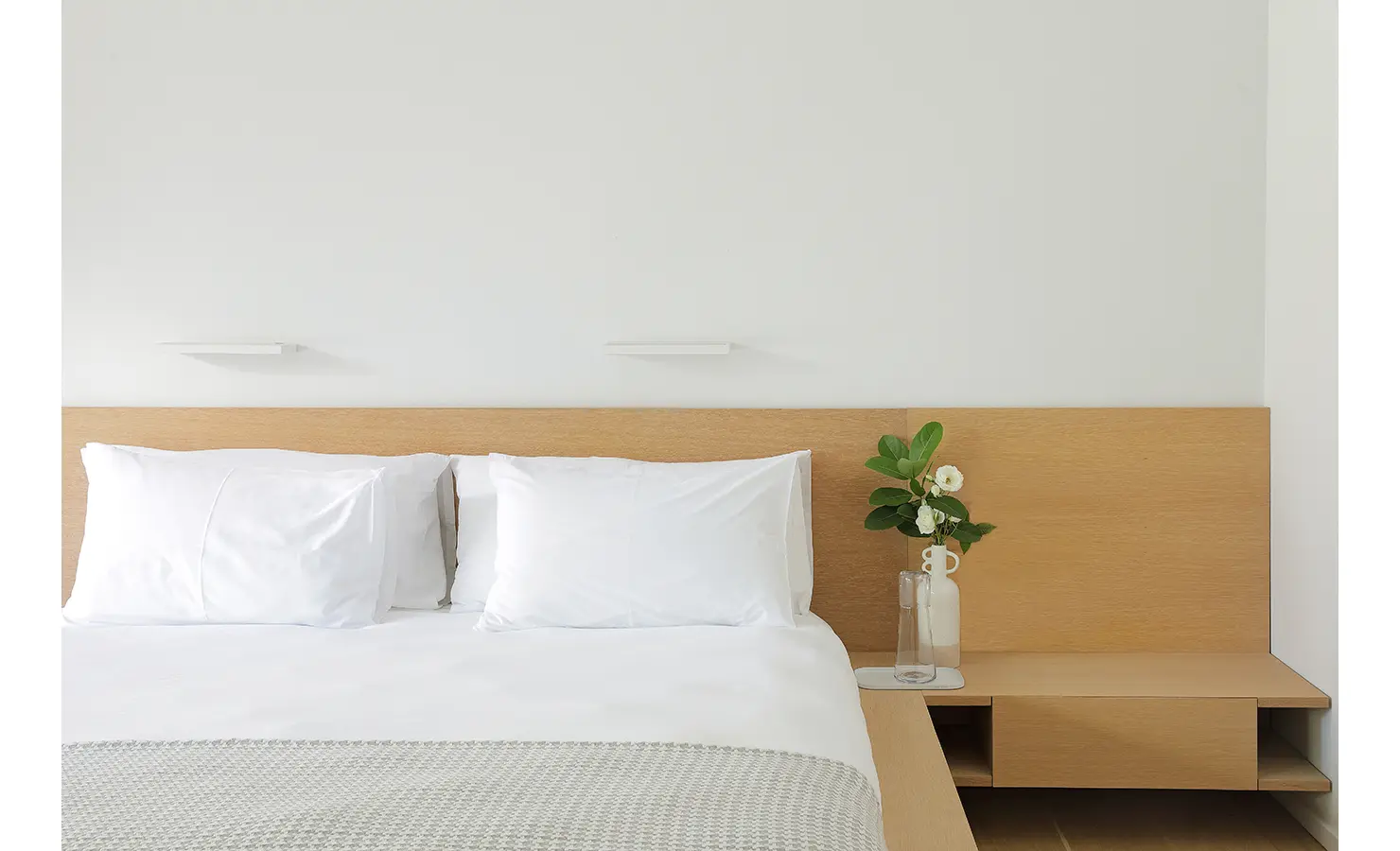 Perfect Hideaways A close-up of a minimalist bed with white pillows, a pale timber headboard, and a simple vase with greenery adding softness to the clean design Pure Living, Bishopscourt