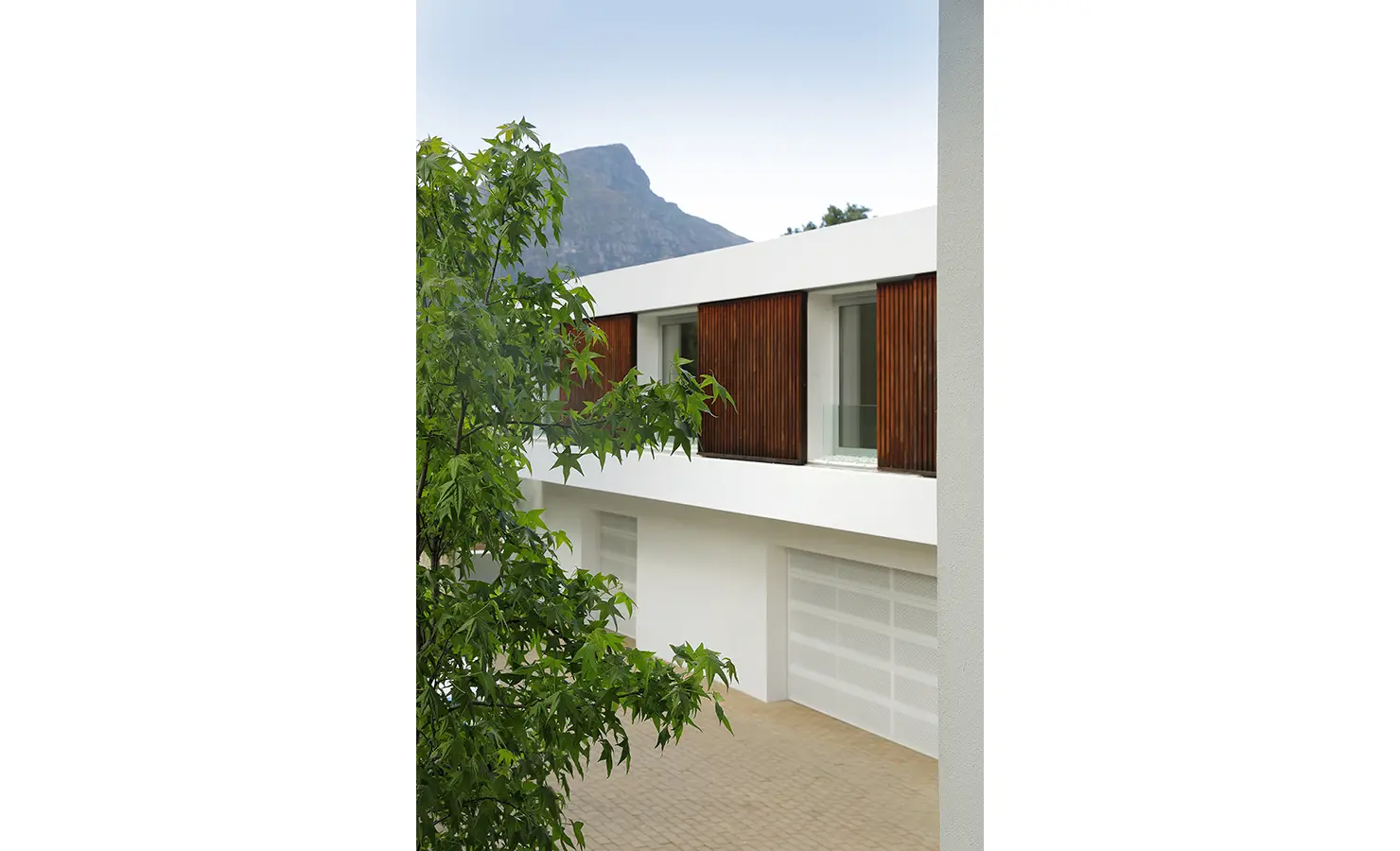 Perfect Hideaways A contemporary façade with timber shutters above a pale brick driveway, framed by leafy trees and mountain views in the distance Pure Living, Bishopscourt