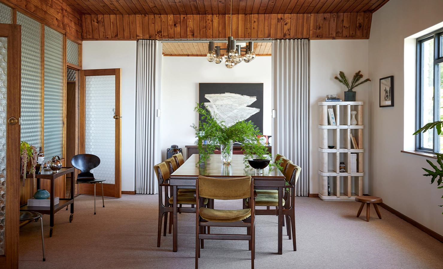 Perfect Hideaways a warm, mid-century–inspired dining room featuring a wooden table surrounded by vintage upholstered chairs, a lush green arrangement centered on the table, textured glass doors, wood-paneled ceiling, sculptural shelving, and soft natural light creating a serene, curated atmosphere Camps Bay, The Liminal