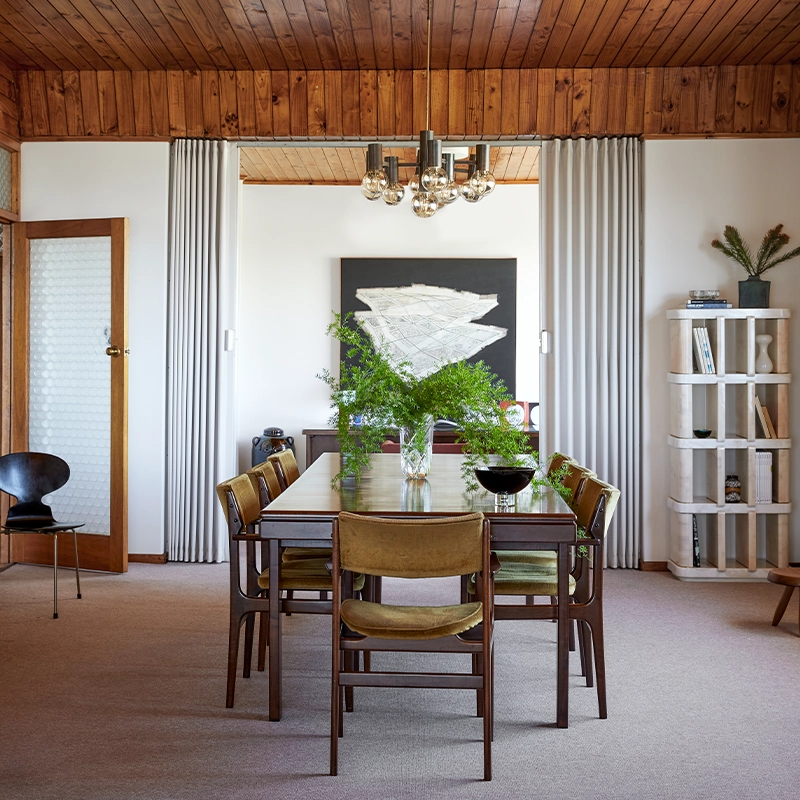 Perfect Hideaways a warm, mid-century–inspired dining room featuring a wooden table surrounded by vintage upholstered chairs, a lush green arrangement centered on the table, textured glass doors, wood-paneled ceiling, sculptural shelving, and soft natural light creating a serene, curated atmosphere Camps Bay, The Liminal