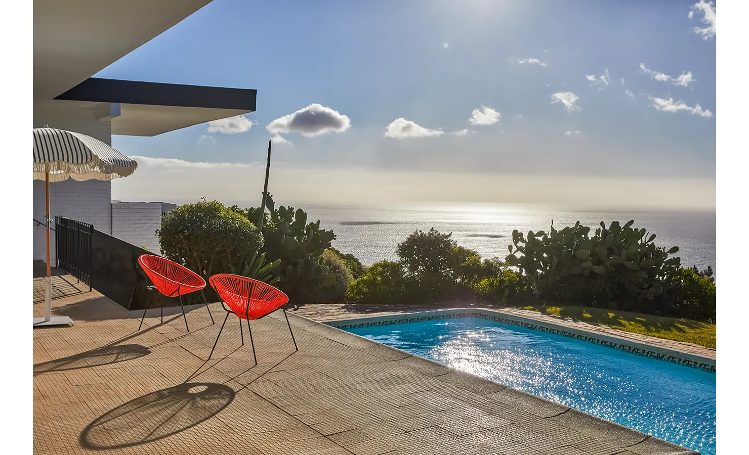Perfect Hideaways a sunlit pool terrace overlooking the ocean, featuring two bright red modern chairs casting long shadows on the tiled patio beside a shimmering blue pool and lush coastal greenery under a soft, hazy sky Camps Bay, The Liminal